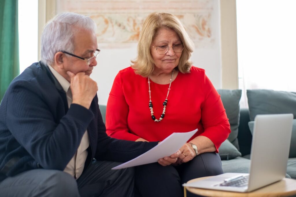 Older couple reviewing insurance documents together while sitting on the couch with a laptop open in front of them